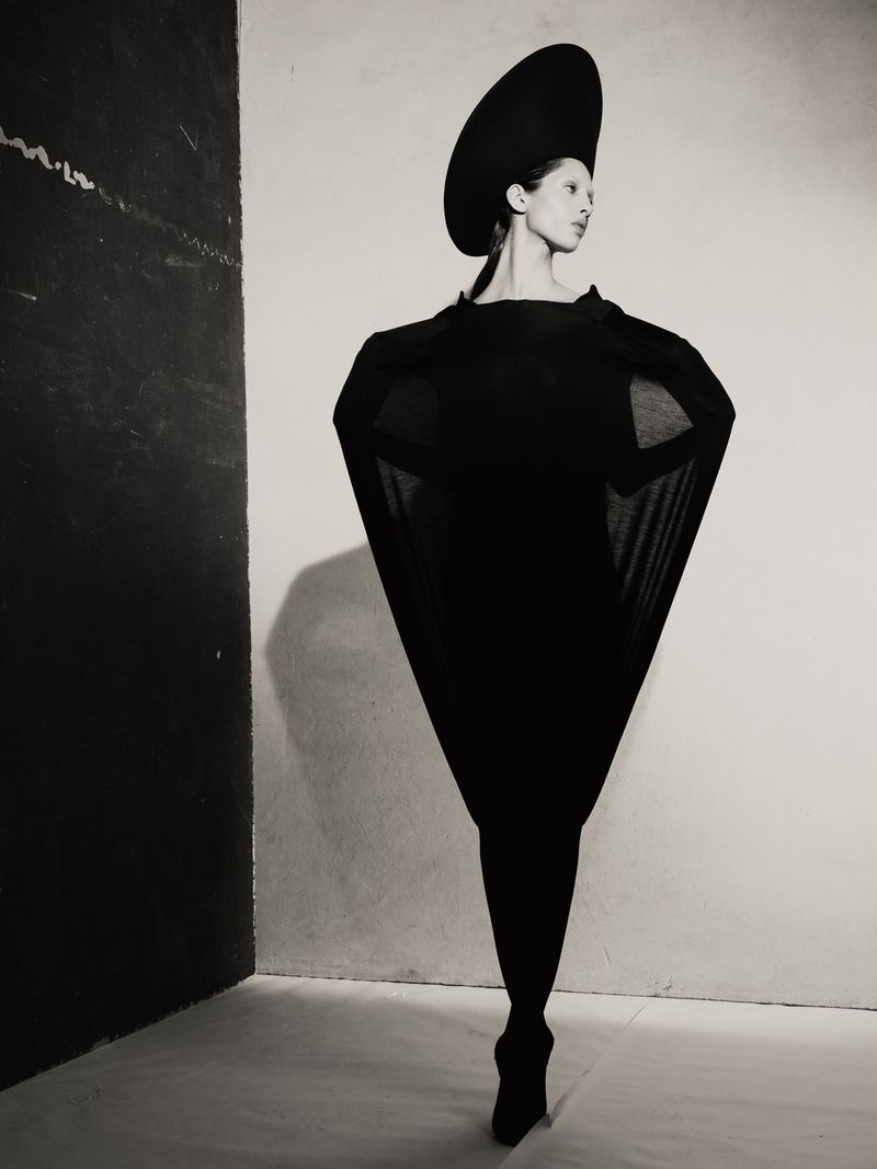 Fashion photo of a woman in a sculptural black dress and hat, posing in a studio with dark and light walls. Monochromatic, elegant.