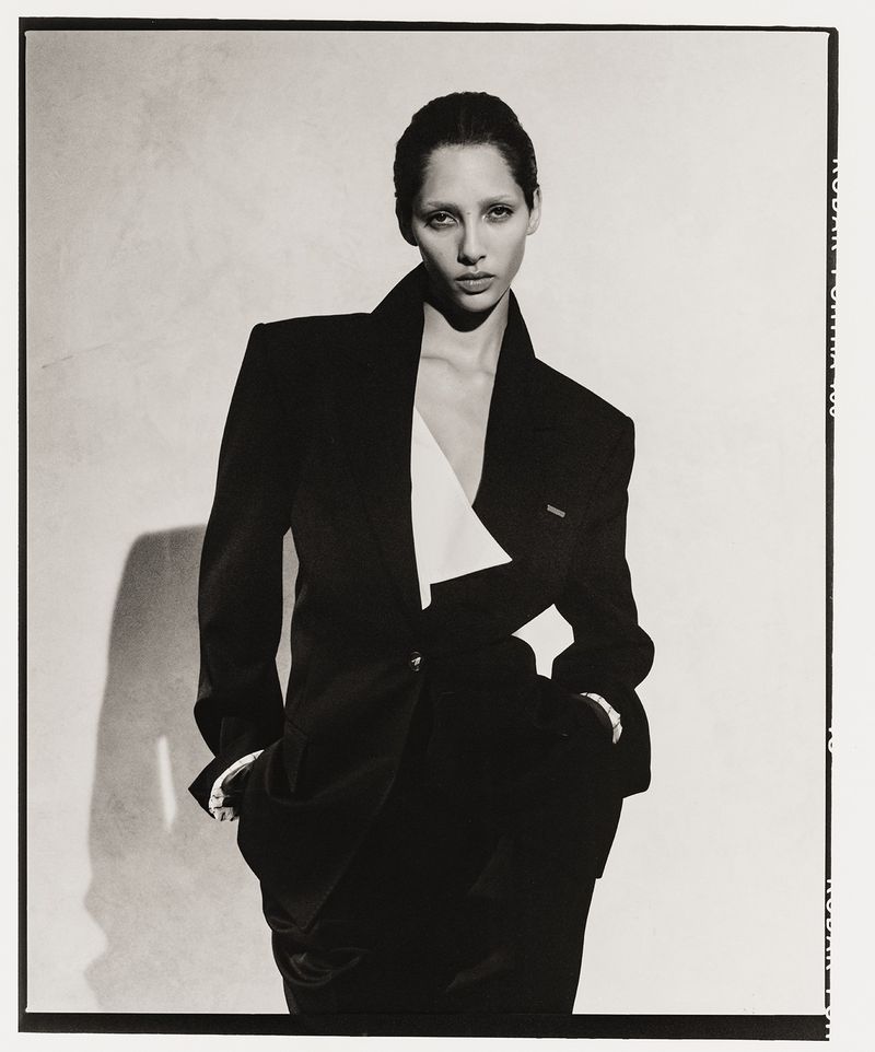A striking studio portrait of a young adult woman wearing a black blazer with white lapel, set against a soft, neutral background.