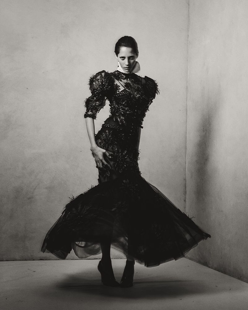 Monochrome fashion portrait of a woman in an ornate black dress with puffed sleeves, standing in a corner, captured in a studio setting.