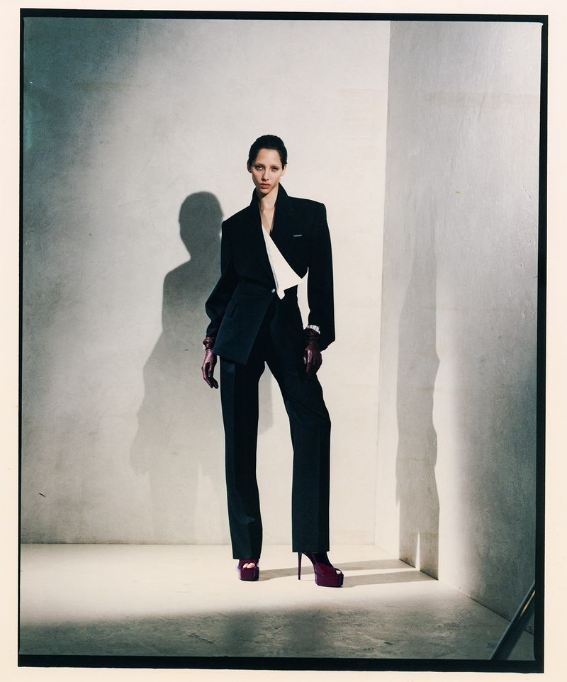 Model Yasmin Wijnaldum wearing a black pantsuit and platform heels standing against a white studio corner wall.