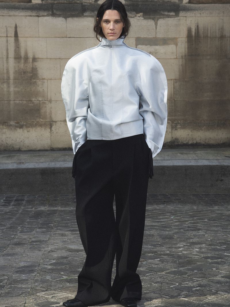 Fashion model poses in a light blue jacket with large shoulder pads and black pants on a stone surface in front of a stone wall.