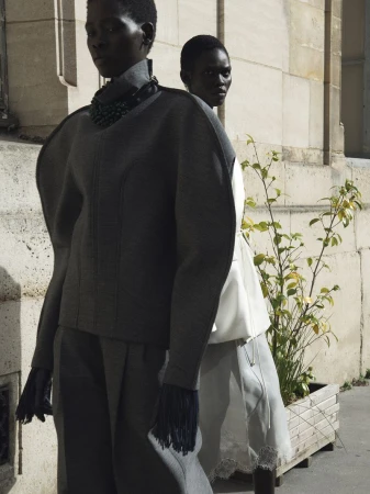 Fashion shot of two dark-skinned female models wearing gray wool and white outfits, posed against a stone building and greenery.