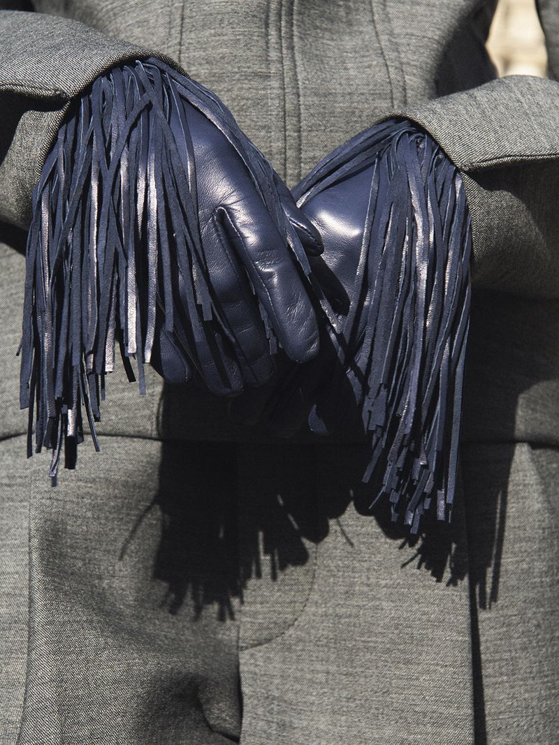 Dark blue leather gloves with long fringe on a grey wool coat. Fashion accessory for cold weather and protection with visible texture.
