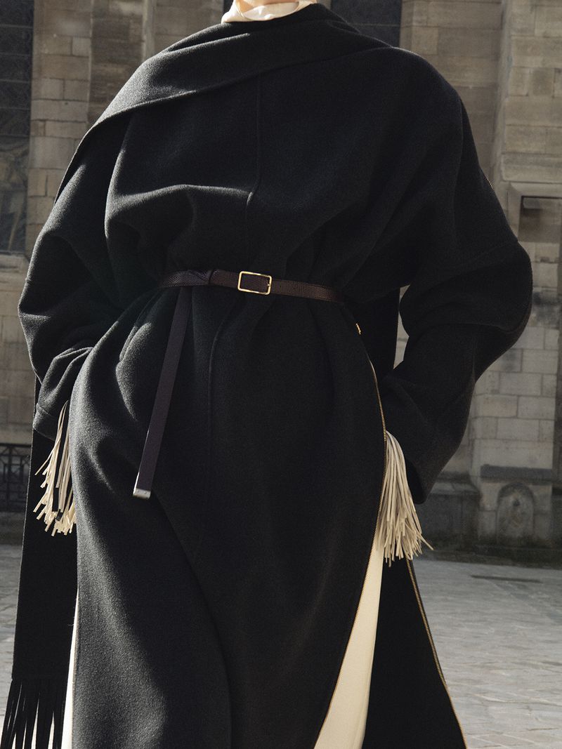 Model wearing a structured black coat with fringed hem and thin belt against a stone building exterior