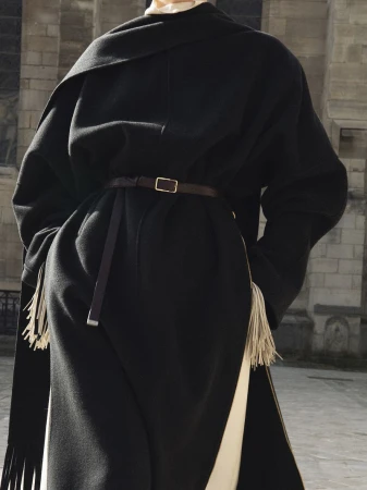 Model wearing a structured black coat with fringed hem and thin belt against a stone building exterior