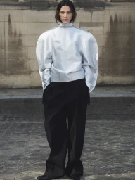 Model wearing a pale structured jacket with voluminous sleeves and dark trousers standing against an old stone wall