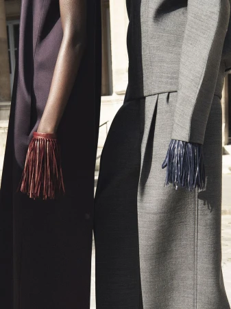 Close-up of two models in structured garments wearing fringed leather bracelets outdoors in the daylight.
