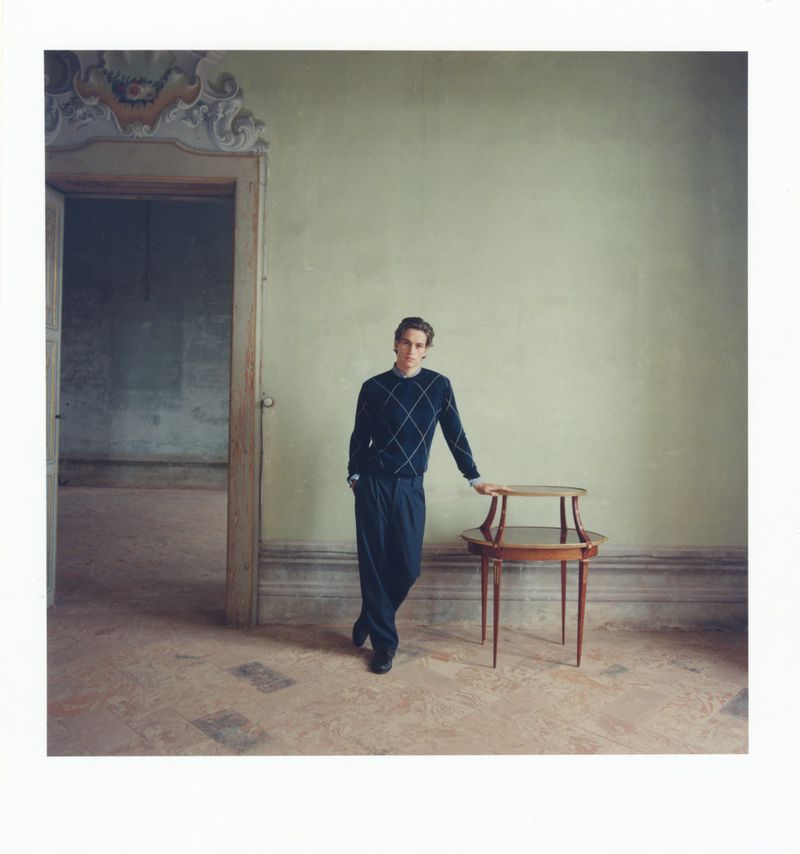 Elegant man in navy argyle sweater and dark trousers poses next to a wooden side table in a spacious, ornate room with green walls.