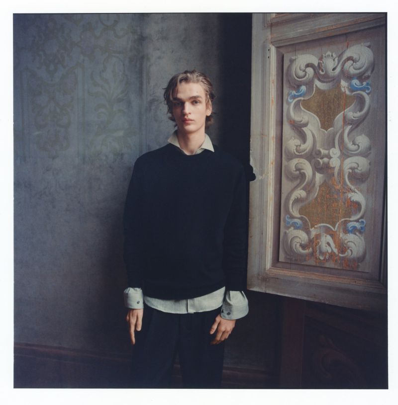 Young man in dark sweater and collared shirt poses near an ornately carved and painted door indoors against patterned wallpaper.