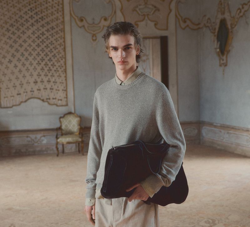 Fashion portrait of a young adult male model in a grey sweater holding a large dark bag in an ornate interior space.