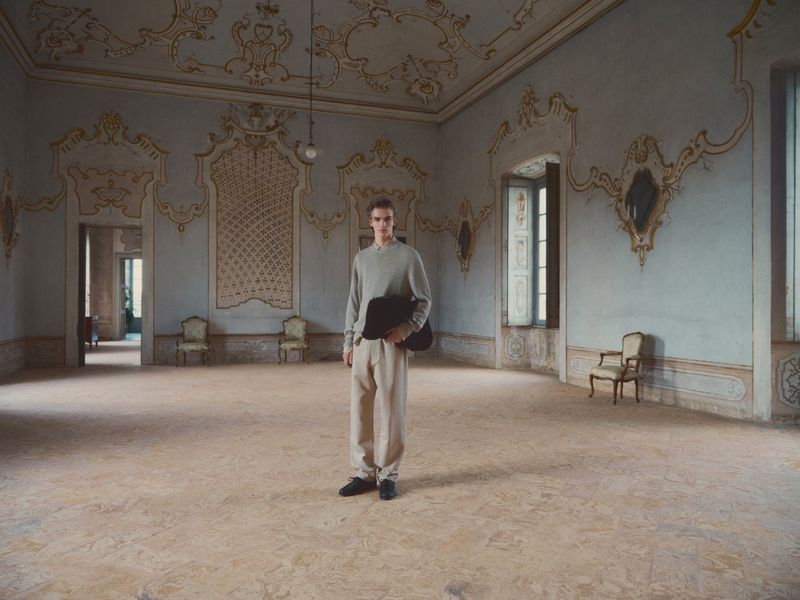 Young man stands in the center of an ornate, light blue and gold room with high ceilings, holding a laptop, and wearing casual clothes.