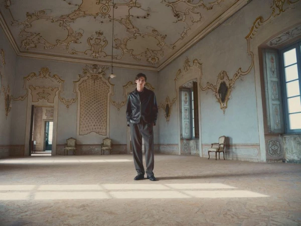 Full-length portrait of a young man in a black jacket and gray pants standing in a large empty room with ornate architectural details.