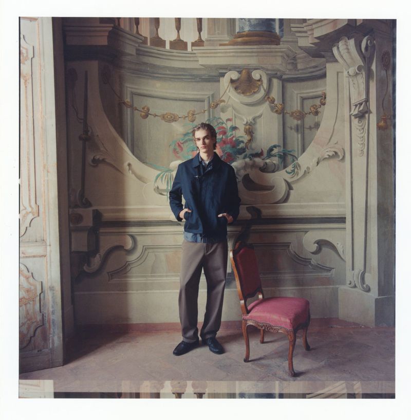 Fashion shot of a young adult man in a dark jacket and light pants standing in an ornate, painted interior near a red chair.