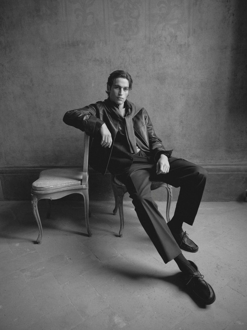 A stylish adult male sits angled on a vintage chair indoors, wearing a black leather jacket and dark pants, against a grey patterned backdrop.