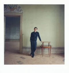 A young male model posing next to a small wooden side table in an elegant room with a pale green wall and an arched doorway.