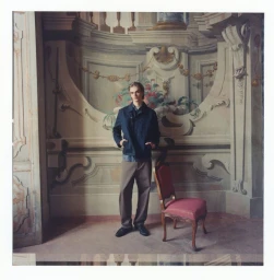 A young male model standing indoors against an ornate painted wall next to a red upholstered vintage chair.