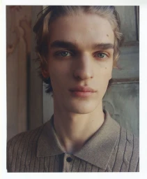 Close-up portrait of a young male model wearing a grey collared polo shirt against a textured architectural background.