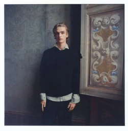 A young male model wearing a dark sweater and collared shirt standing against an ornate wall with a carved wooden panel.