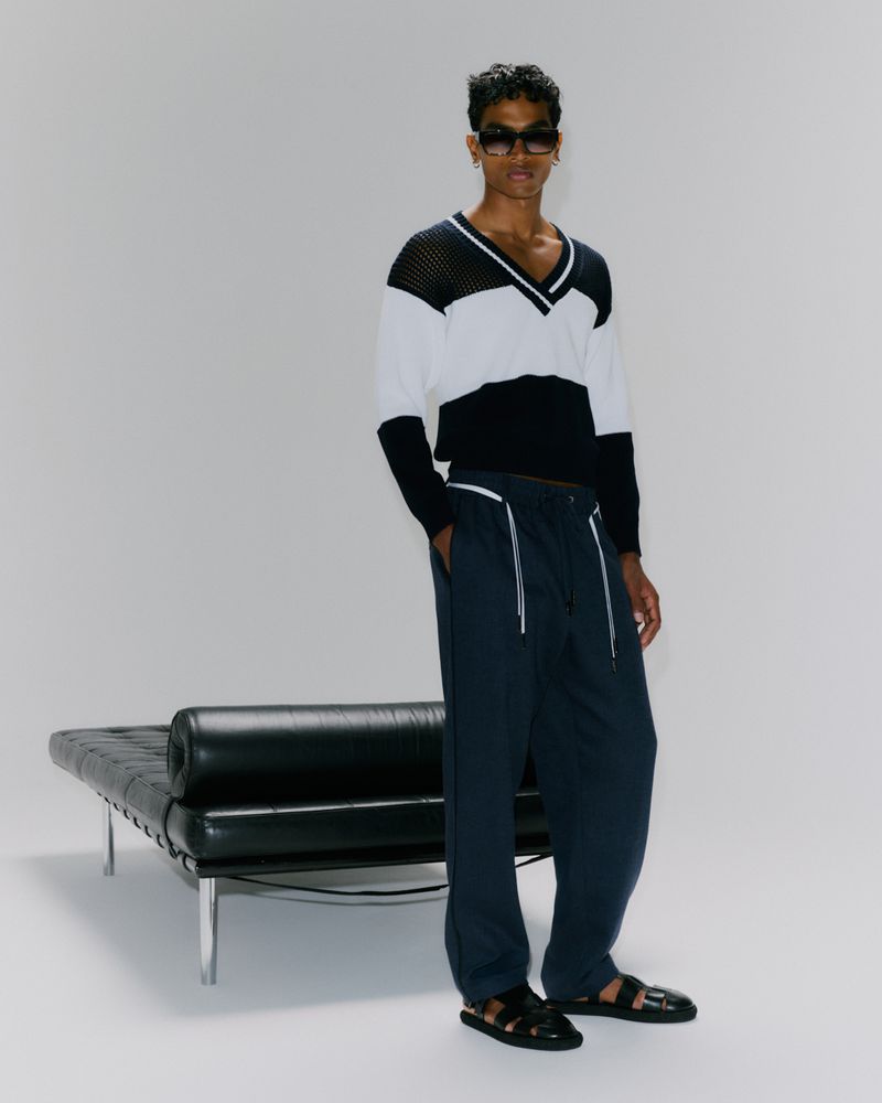 Adult male in a white, black, and blue v-neck sweater with blue pants and black sandals, posed in a studio setting, with a black bench.