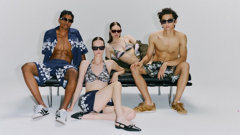 Group of four fashion models wearing swimwear and floral patterned casual wear sitting in front of a white wall in a studio.