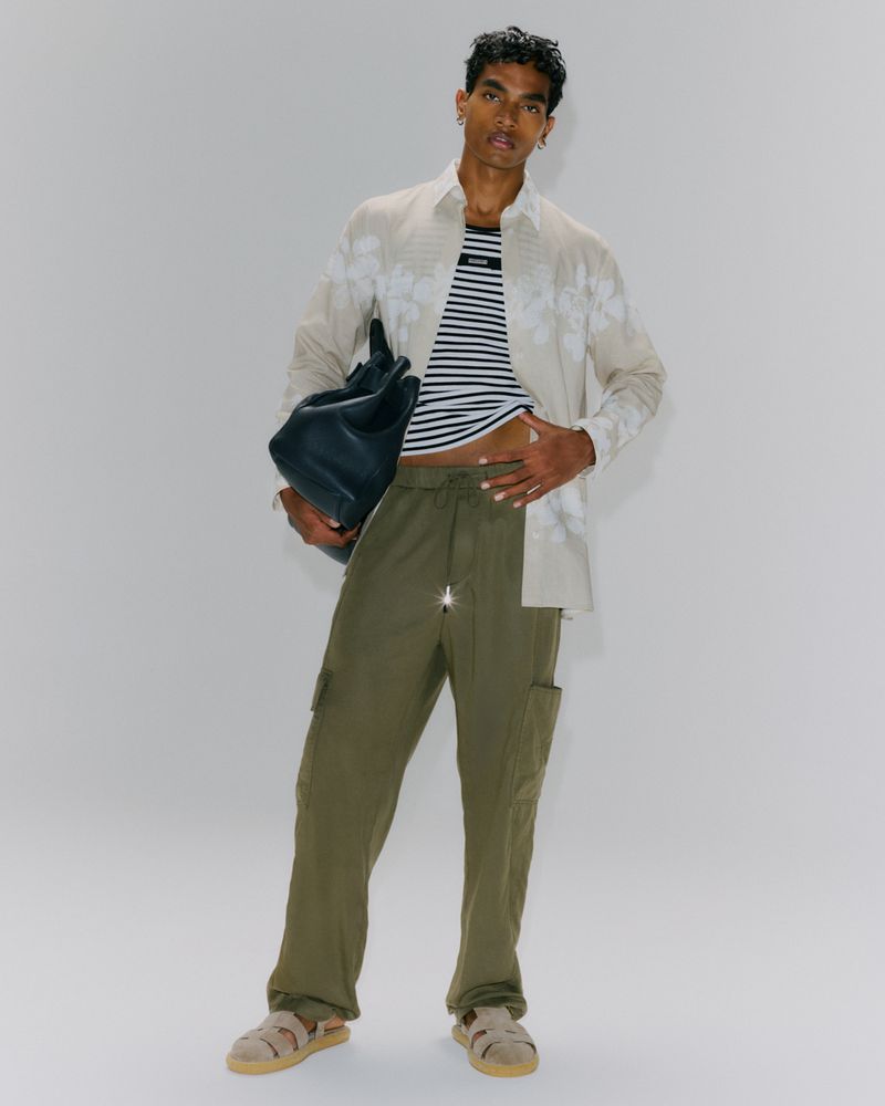 Adult male poses indoors wearing striped t-shirt, floral shirt, and olive cargo pants while holding a dark-colored leather bag.