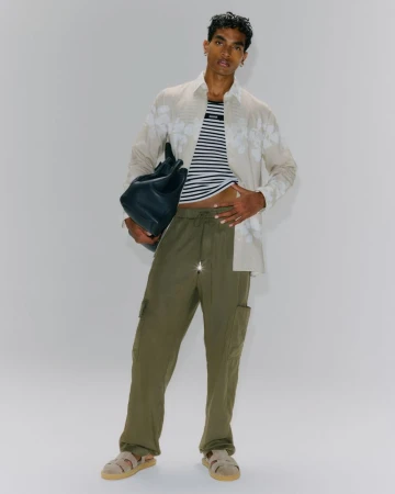 Adult male poses indoors wearing striped t-shirt, floral shirt, and olive cargo pants while holding a dark-colored leather bag.
