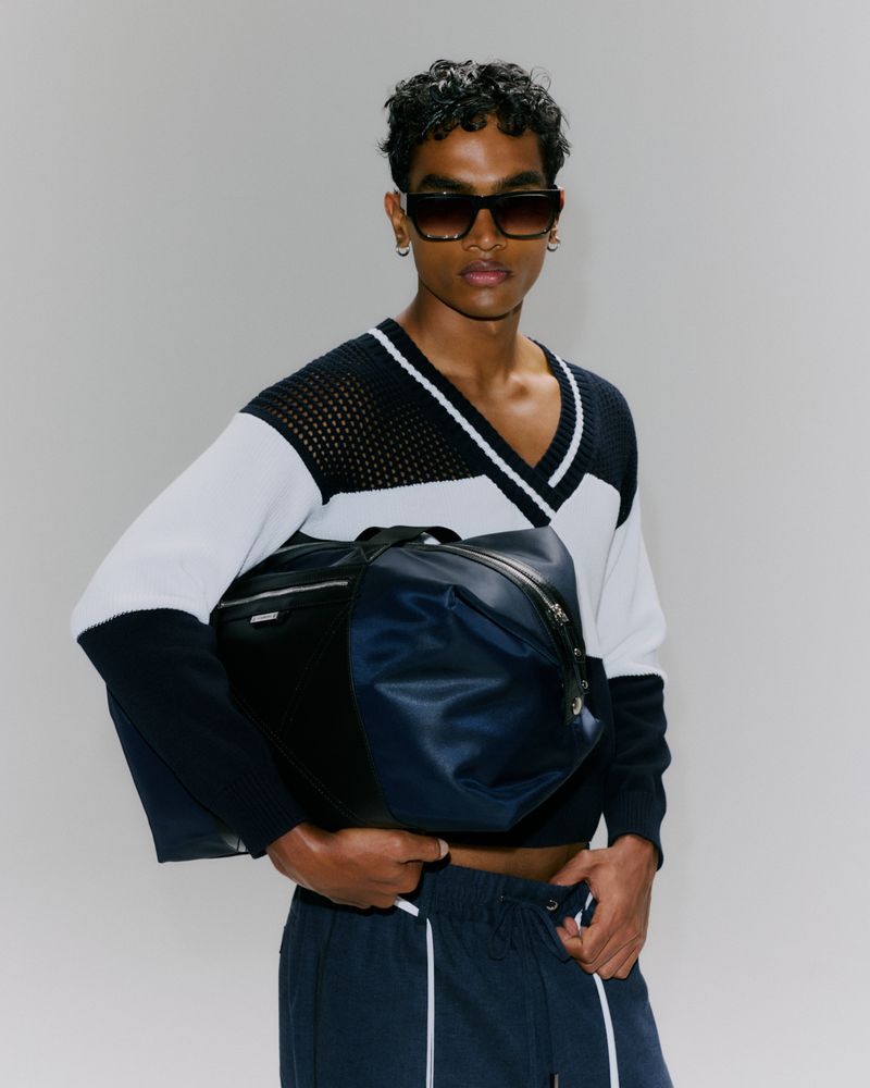 A young adult male poses indoors wearing a black and white sweater, navy sweatpants, and sunglasses, holding a navy duffel bag.