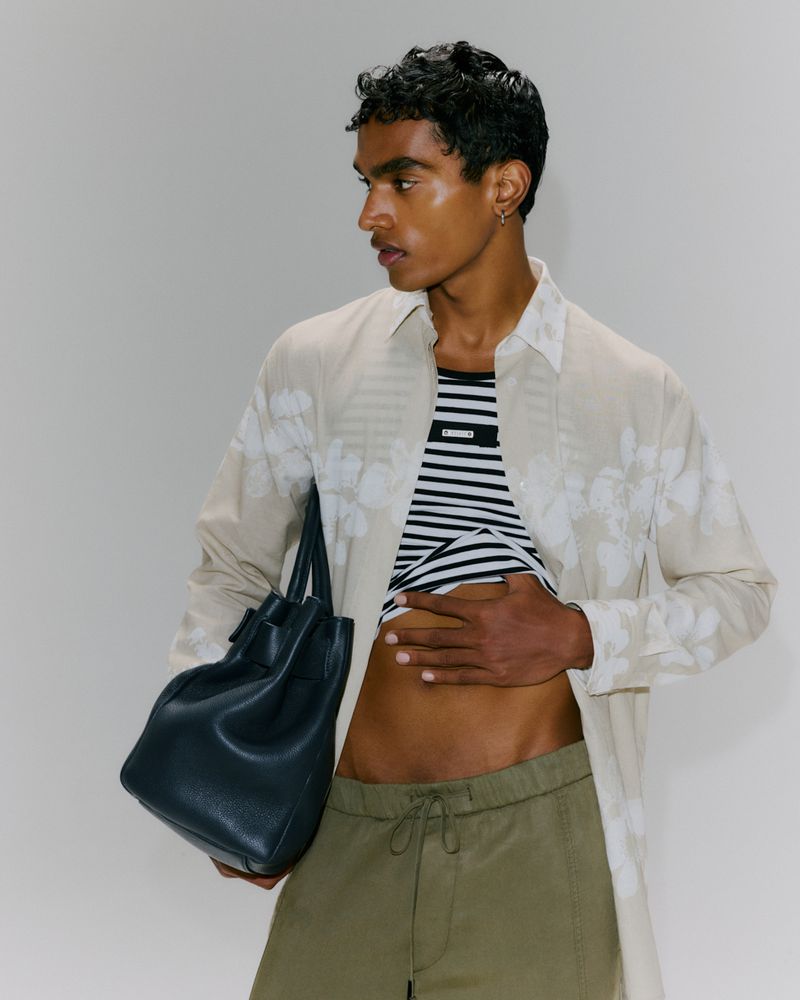 A male model poses in an open floral button-down shirt with a striped top, green trousers, and a blue leather tote bag against a white backdrop.
