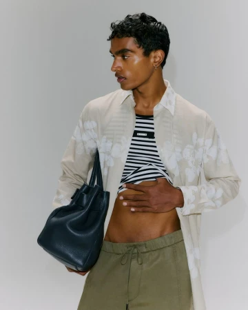 Model wearing a patterned shirt and olive trousers holding a dark leather handbag against a grey backdrop.