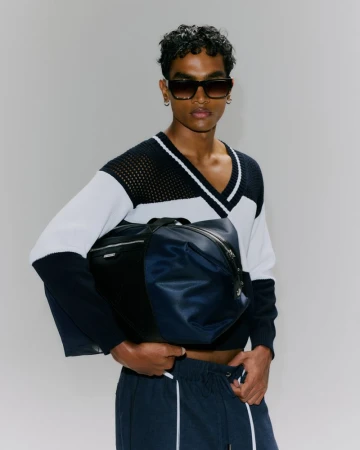 Model wearing dark sunglasses and a color-blocked knit sweater holding a navy duffel bag against a plain light grey backdrop.