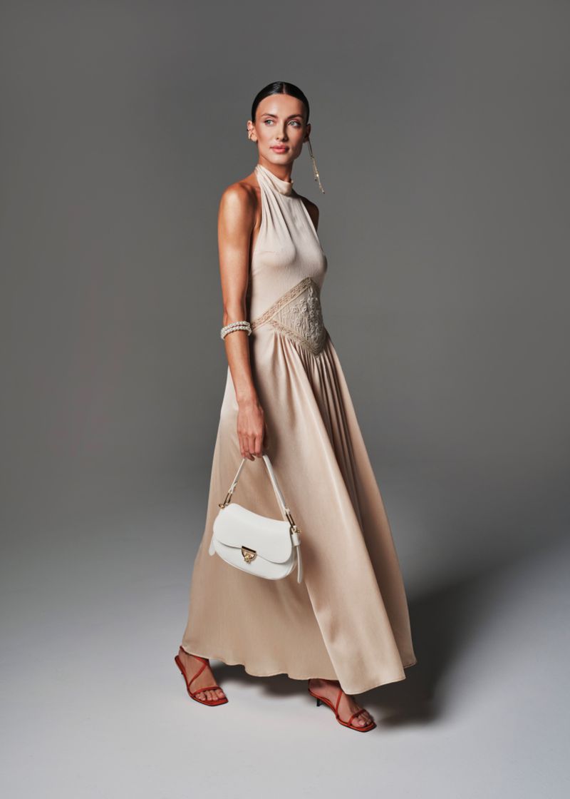 Elegant adult woman wearing a long, tan dress and red sandals carries a white purse in a studio setting on a gray background.