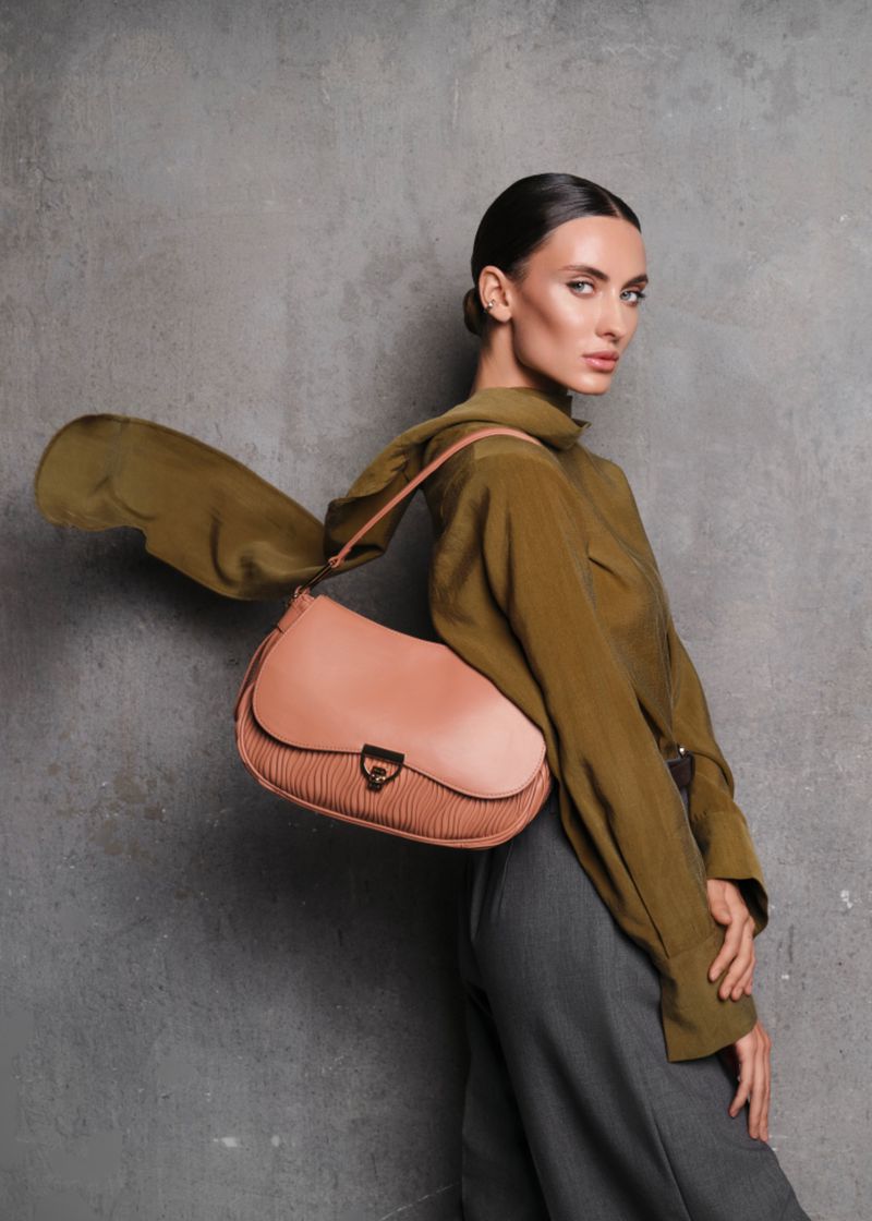 Fashion portrait of a woman wearing an olive green shirt with a salmon-colored purse in front of a textured gray wall.