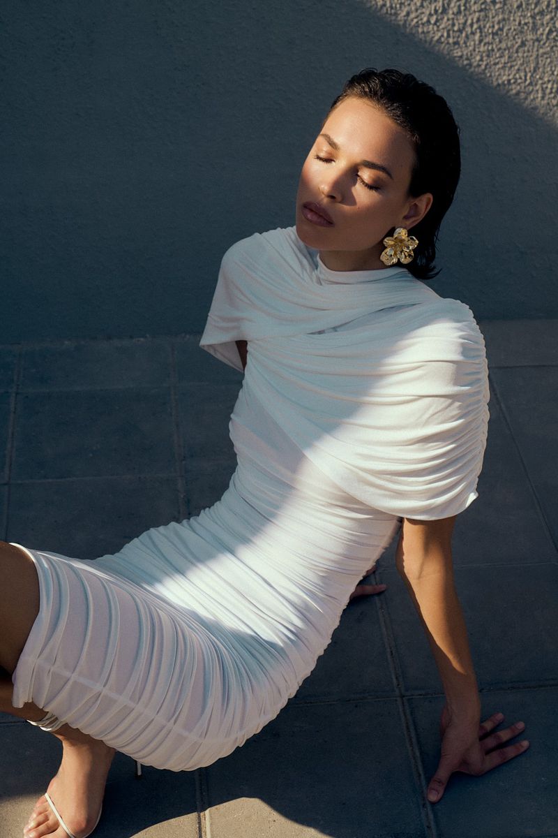 Woman in a draped white dress and gold earrings poses with closed eyes against a textured background, with sunlight and shadows.