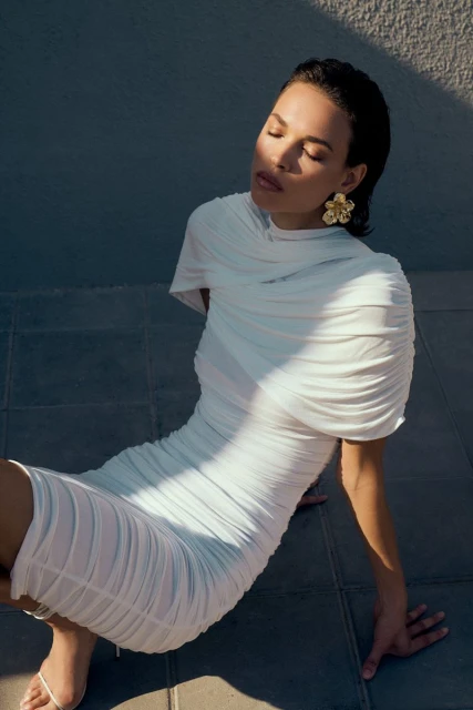 Woman in a draped white dress and gold earrings poses with closed eyes against a textured background, with sunlight and shadows.