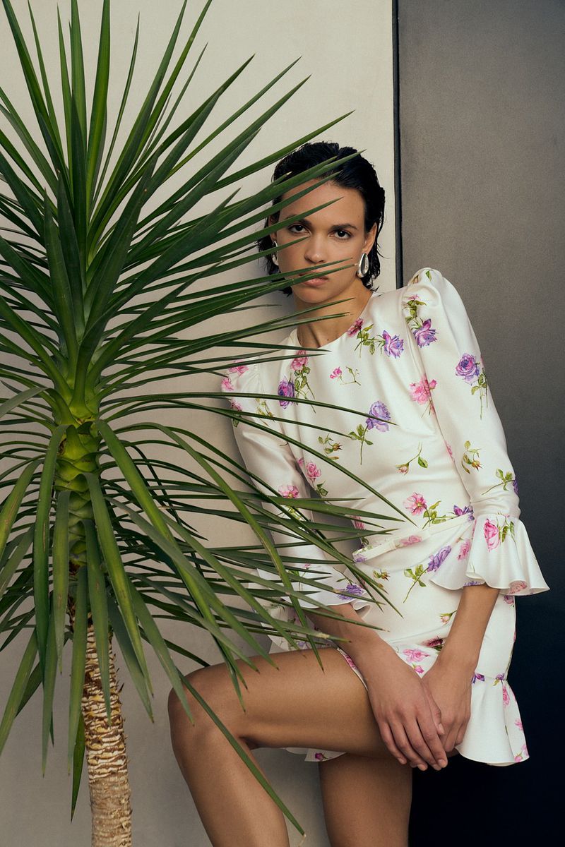 A woman in a floral dress poses next to a plant with long leaves. The image emphasizes fashion and botanical elements in an indoor setting.