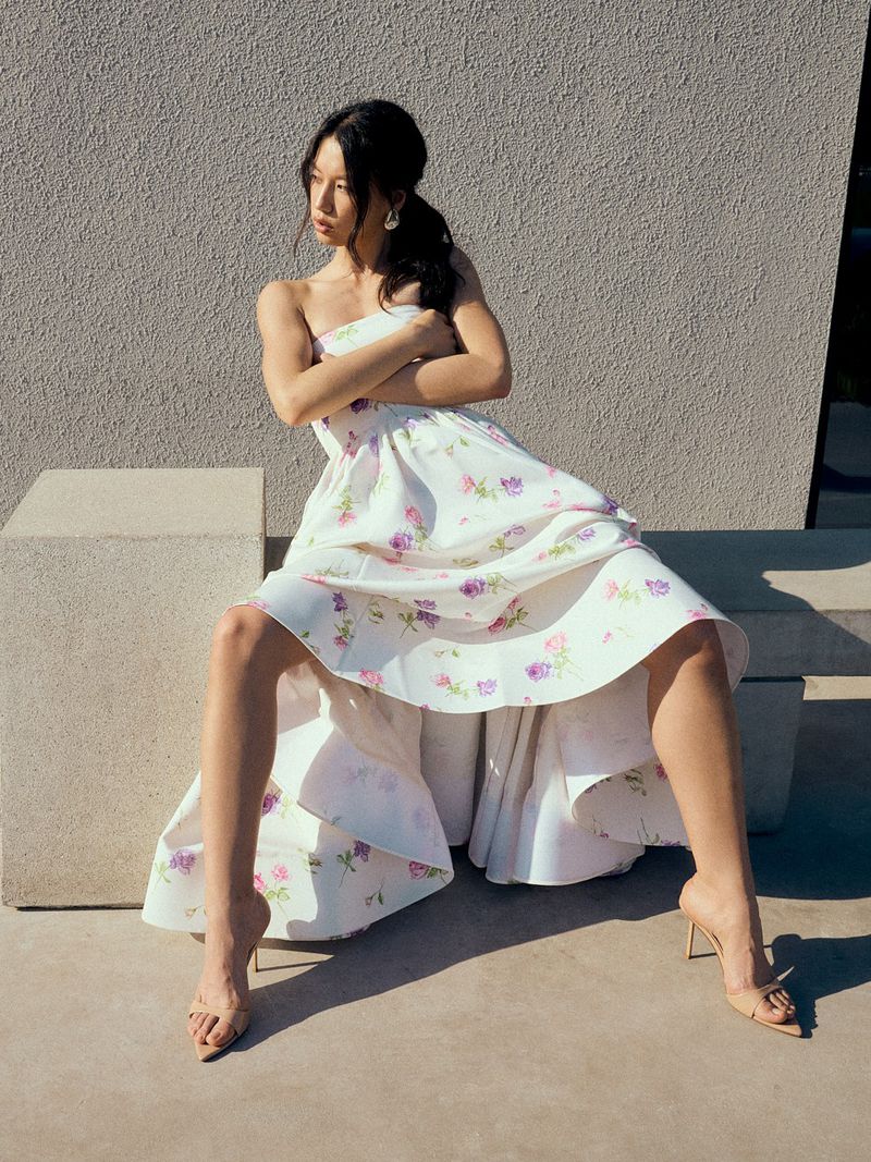 An Asian woman poses outdoors on concrete blocks, wearing a floral strapless dress and high heels in bright sunlight.