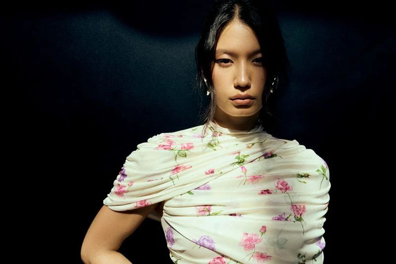 Portrait of an Asian woman with a serious expression, wearing a floral-patterned top against a dark background in a studio shot.