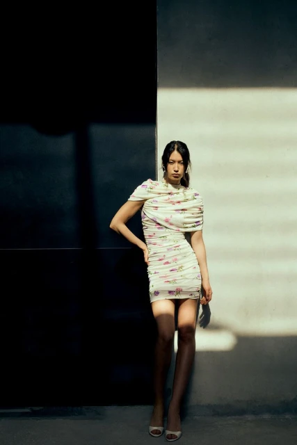 A woman in a floral dress poses against a split black and white wall, illuminated by sunlight creating distinct light and shadow patterns.