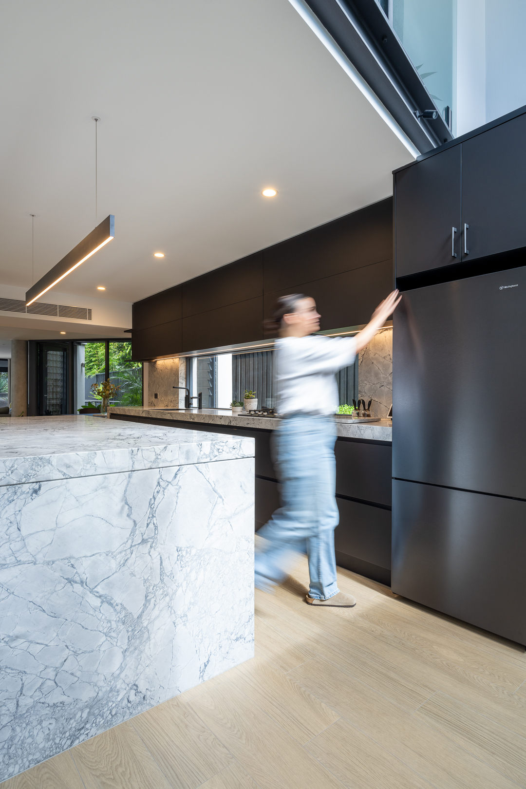 Woman reaching into a sleek, modern, black refrigerator in an open-plan kitchen with marble countertops and light wood floors. 