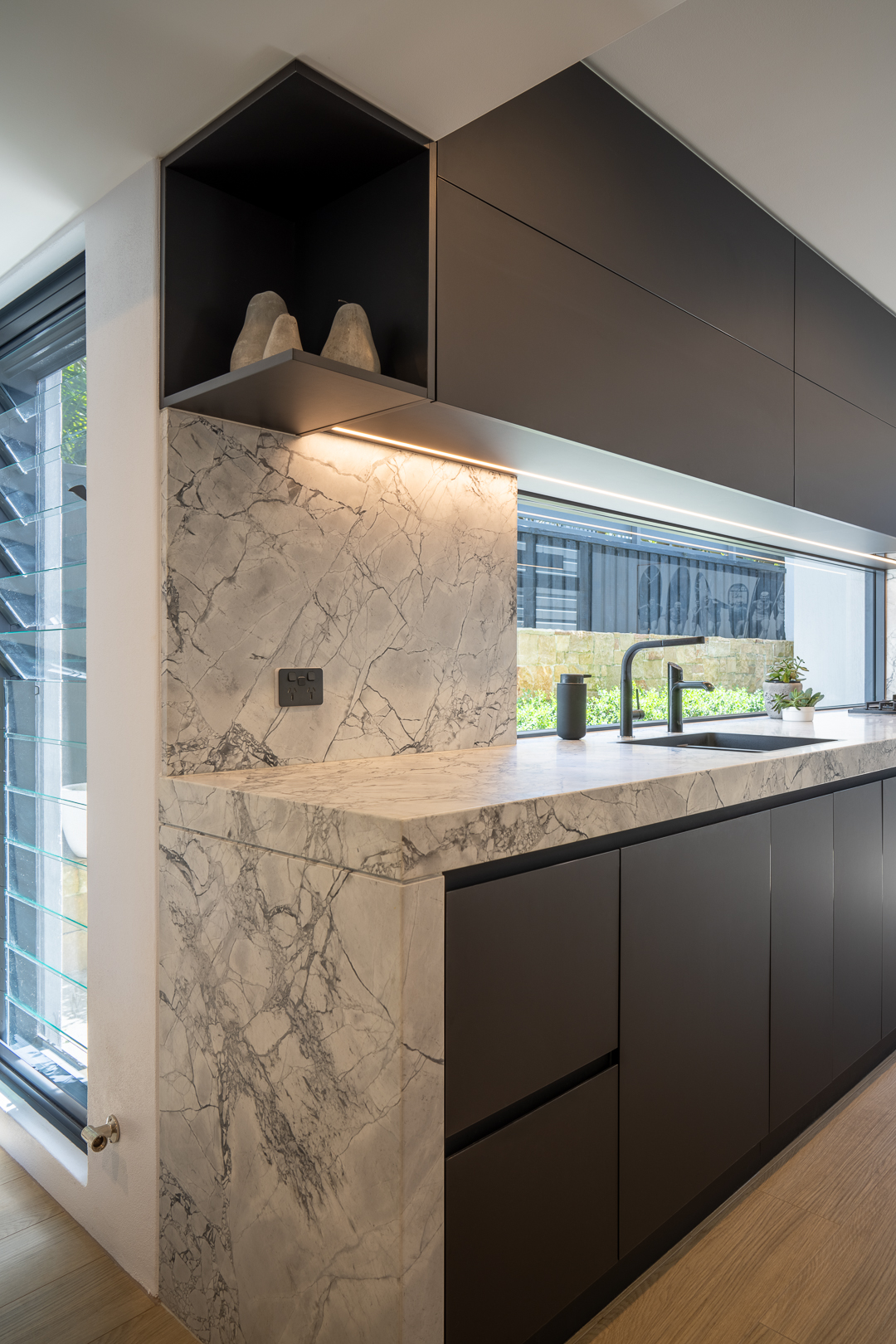 Sleek modern kitchen interior with dark cabinetry, marble countertops and backsplash, featuring decorative objects and ambient lighting.
