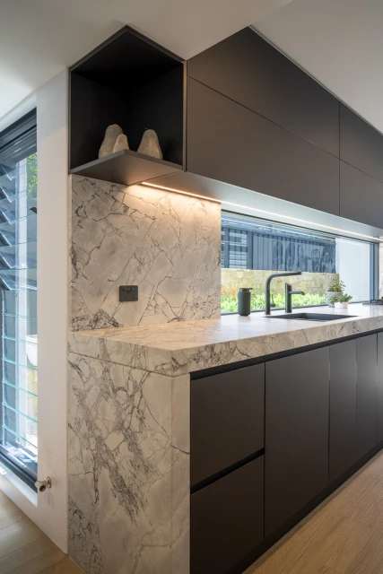 Sleek modern kitchen interior with dark cabinetry, marble countertops and backsplash, featuring decorative objects and ambient lighting.
