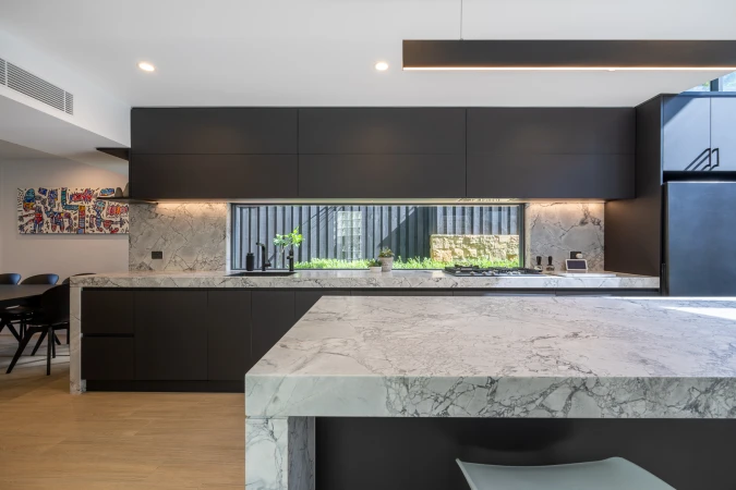 Sleek modern kitchen featuring dark cabinets, marble countertops, and a garden view through a window, with minimalist lighting and decor.