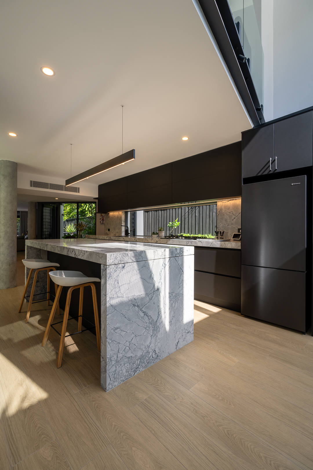 Modern kitchen interior featuring a marble island with stools, dark cabinetry, and hardwood flooring, illuminated by natural light.