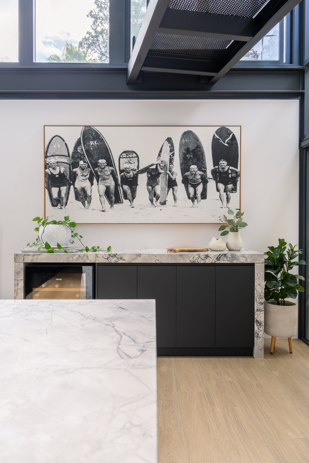 Modern kitchen interior featuring marble countertops, dark cabinets, and an art print of vintage surfers with surfboards on display.