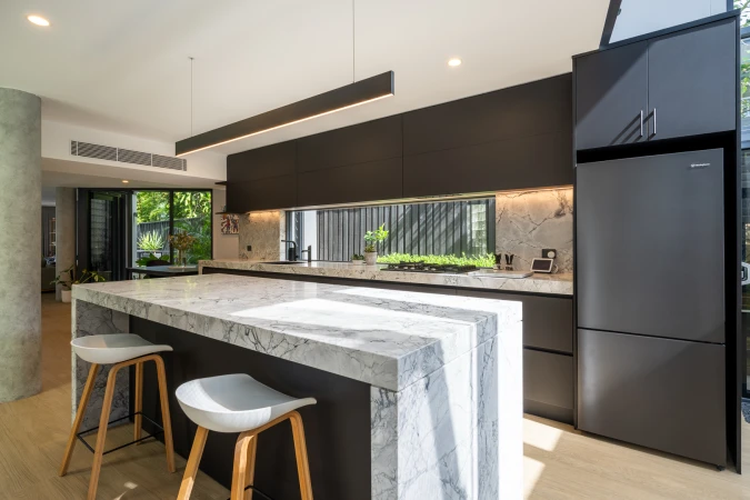 Modern kitchen interior with a marble island, dark cabinets, stainless steel refrigerator, and natural light from outdoor greenery.