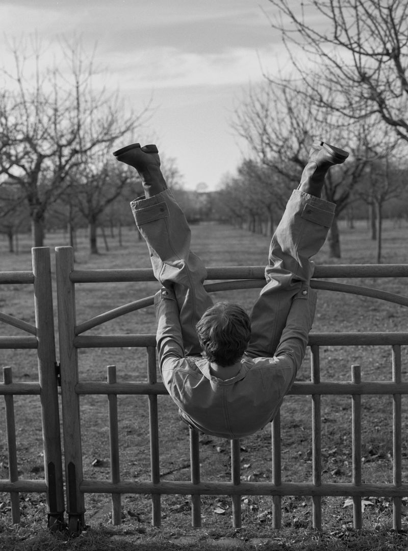 Black and white photo of an adult hanging upside down on a metal fence in a rural setting with trees in the background.