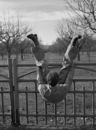 Black and white photo of an adult hanging upside down on a metal fence in a rural setting with trees in the background.
