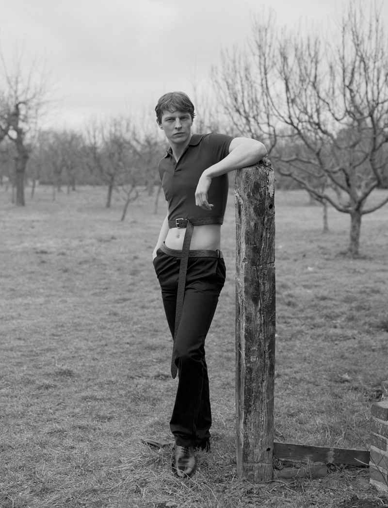 Monochrome portrait of a fair-skinned adult male in polo shirt and trousers leaning against a wooden post in a rural grassy field.