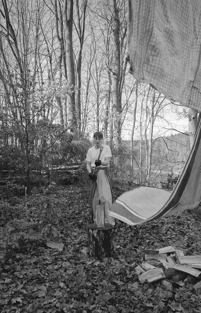 Monochrome outdoor portrait of a young adult standing on a tree stump in a forest holding camera equipment.