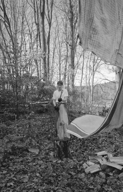 Monochrome outdoor portrait of a young adult standing on a tree stump in a forest holding camera equipment.